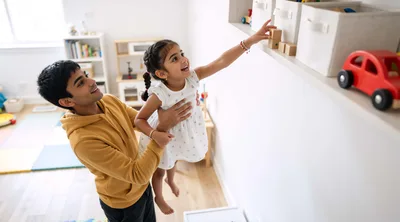 Father Lifting Daughter To Reach Toy On Shelf