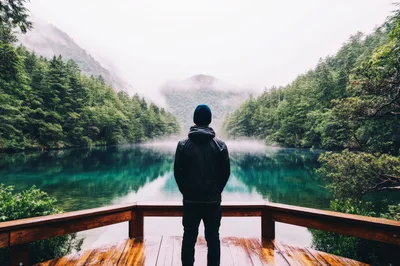 Man Standing On Wooden Deck Overlooking Misty Mountain Lake