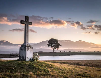 Stone Cross Monument Overlooking A Serene Lake At Sunset