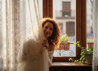 Young Woman Peeking Through Lace Curtains By A Window