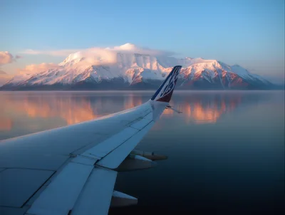 Airplane Wing View Over Snowy Mountains And Calm Water