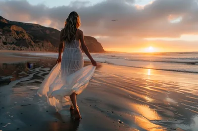 Woman Walking Along Beach At Sunset In White Dress