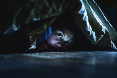 Scared Child Hiding Under Bed Covers In Dark Room