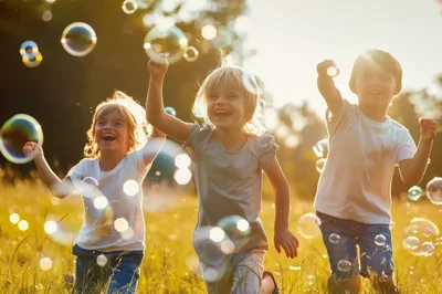 Three Happy Children Running Through Bubbles In A Meadow