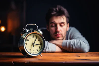 Tired Man Resting Head On Table With Alarm Clock