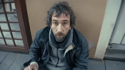 Homeless Man Sitting On Sidewalk Looking Up At Camera