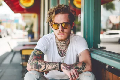 Young Man With Tattoos And Sunglasses Sitting Outdoors