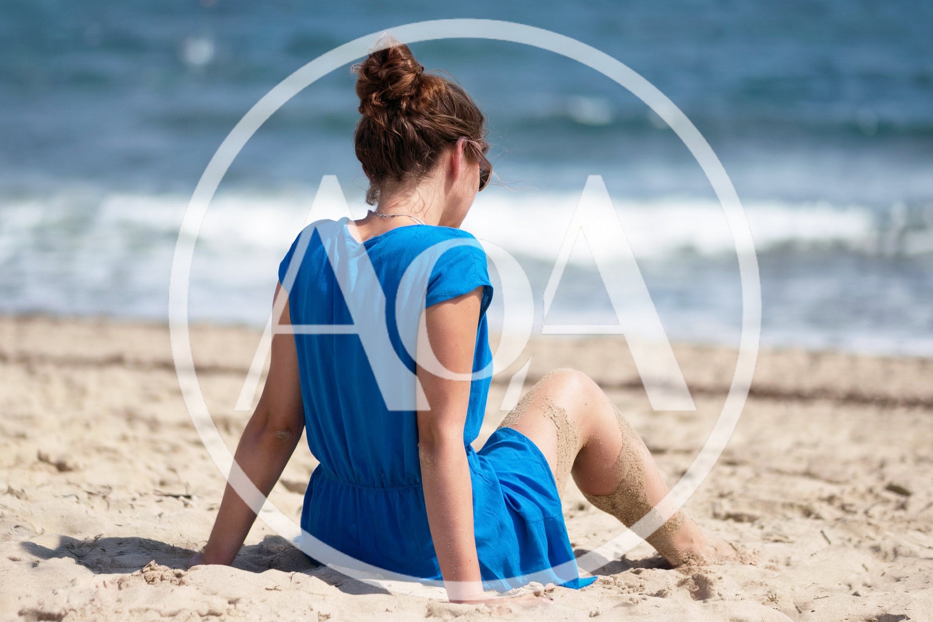 Young Woman Sitting On Sandy Beach In Blue Dress