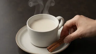 Hand Placing Cinnamon Sticks Beside Steaming Cup Of Milk