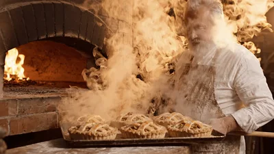 Elderly Baker Removing Fresh Pies From Wood Fired Oven
