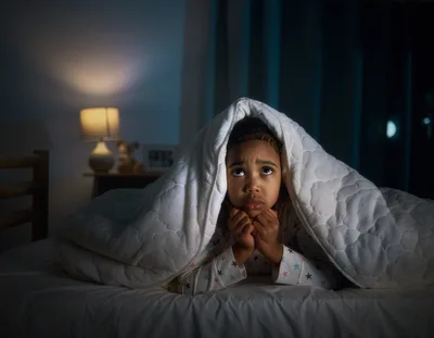 A child is lying in bed at home in dim lighting, peeking out from under the blanket, looking frightened. Night terror concept.