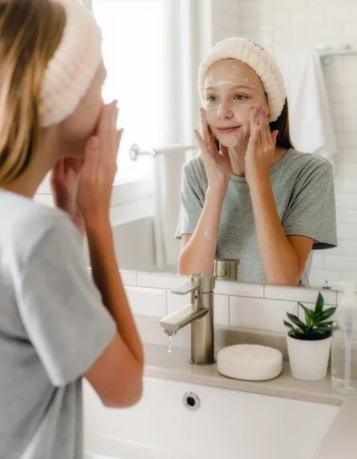 A teenage girl cleans her face with a facial cleanser in front of a mirror in a bright bathroom. Her face is lightly covered in foam, and she is wearing a soft headband.