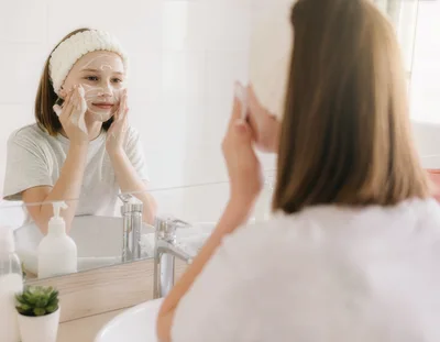 A teenage girl cleans her face with a facial cleanser in front of a mirror in a bright bathroom. Her face is lightly covered in foam, and she is wearing a soft headband.