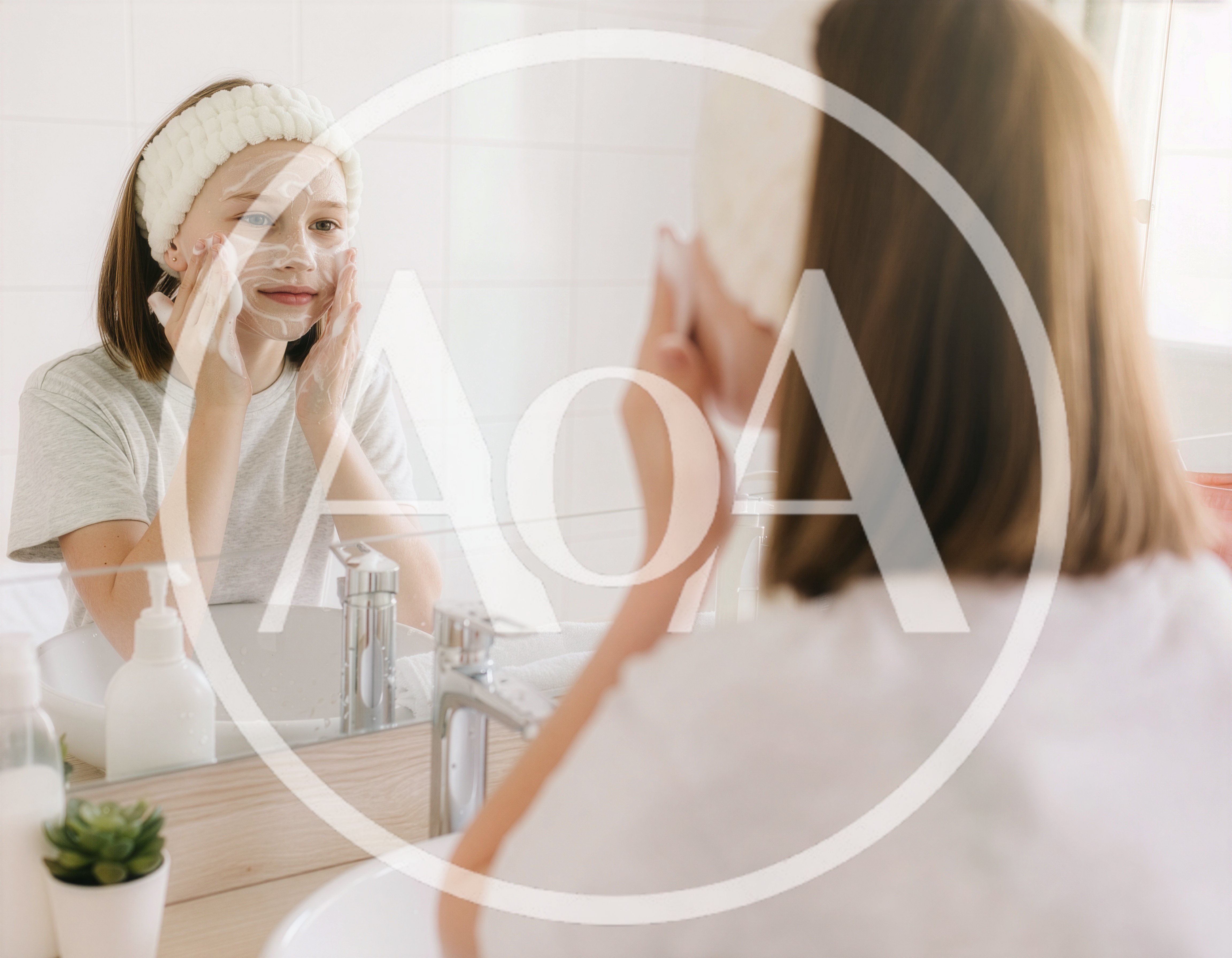 A teenage girl cleans her face with a facial cleanser in front of a mirror in a bright bathroom. Her face is lightly covered in foam, and she is wearing a soft headband.