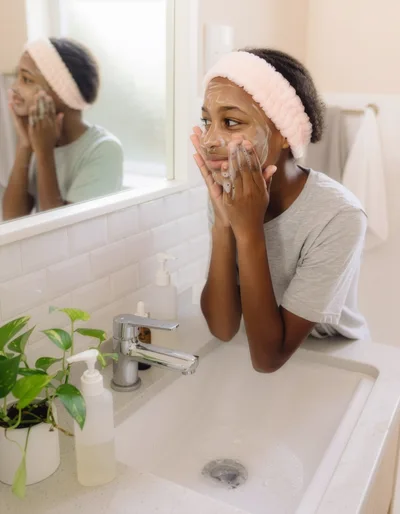 A teenage girl cleans her face with a facial cleanser in front of a mirror in a bright bathroom. Her face is lightly covered in foam, and she is wearing a soft headband.