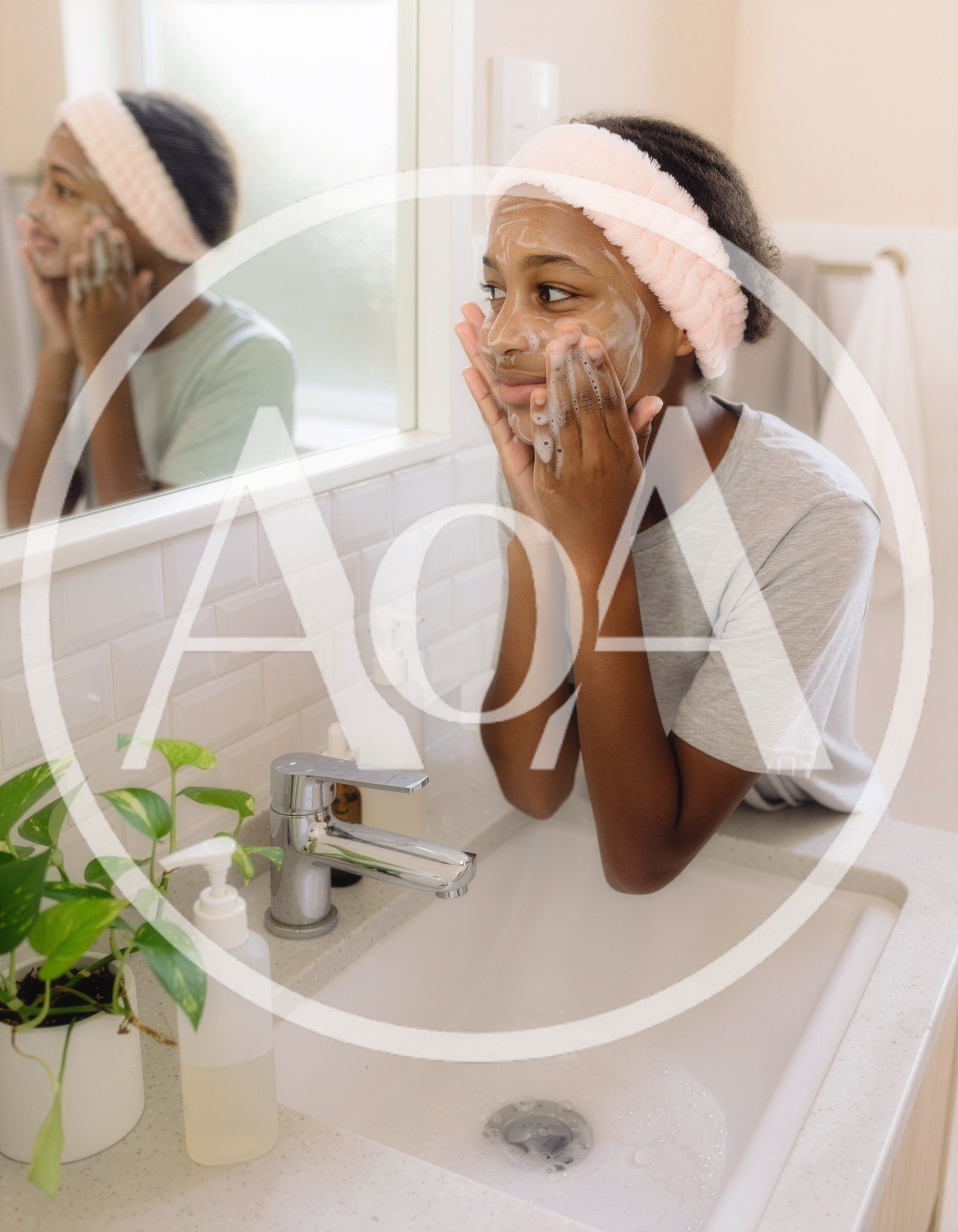 A teenage girl cleans her face with a facial cleanser in front of a mirror in a bright bathroom. Her face is lightly covered in foam, and she is wearing a soft headband.