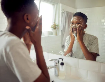 Young African American Boy Washing Face In Bathroom Mirror