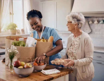Caregiver Helping Elderly Woman Unpack Groceries In Kitchen