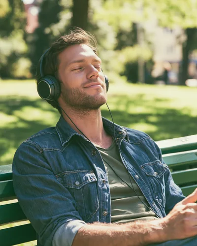 Young Man Relaxing On Park Bench Listening To Music